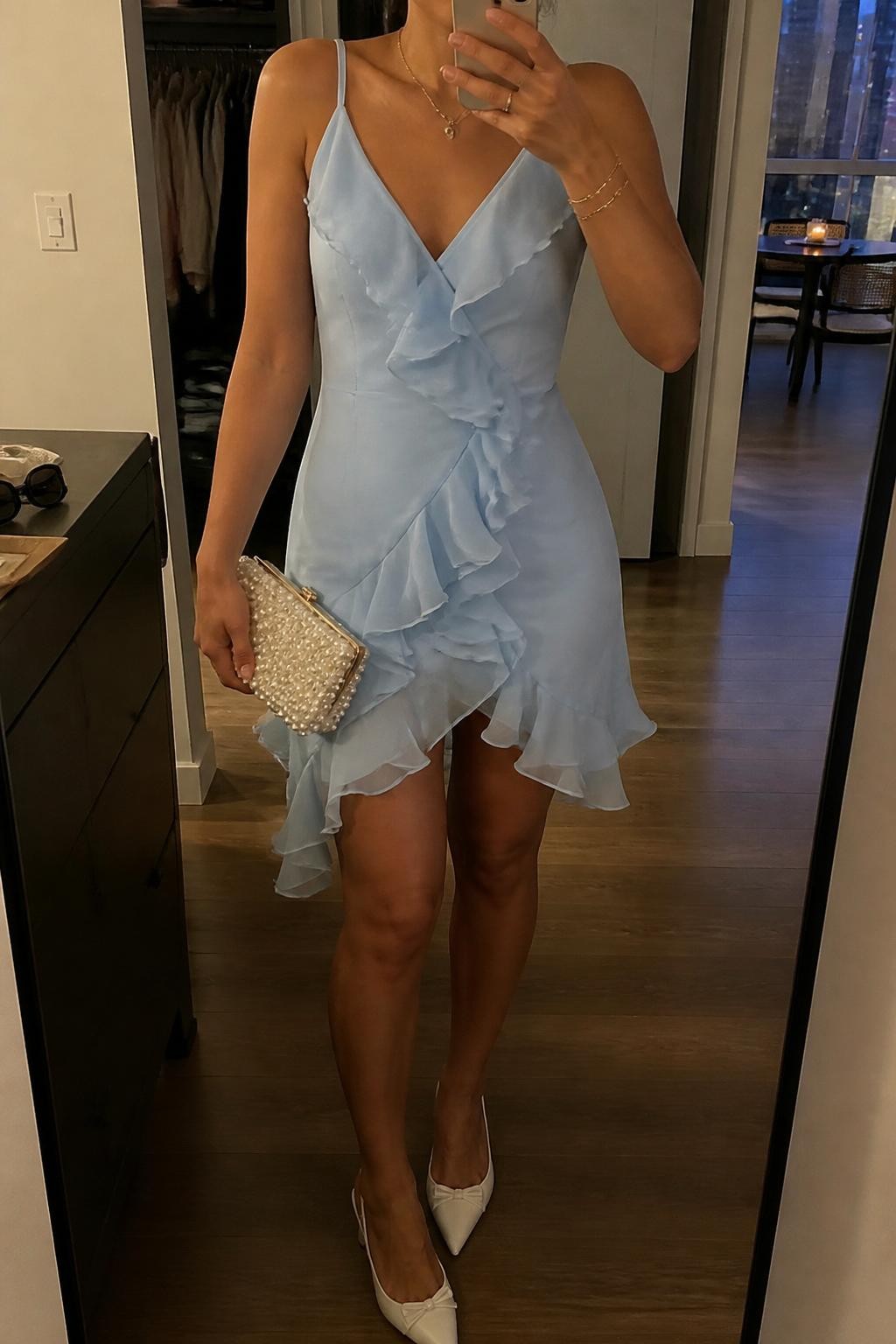 Image prompt
mirror selfie of a woman wearing a sky blue ruffle chiffon dress with white kitten heels, a pearl clutch, and delicate gold jewelry, holding a phone in front of a mirror, cropped at NECK so face doesn't show, with a natural casual selfie pose, in a city apartment, warm moody lighting, including small lived-in details like an open closet behind and sunglasses on a dresser, latest iPhone photo quality.
