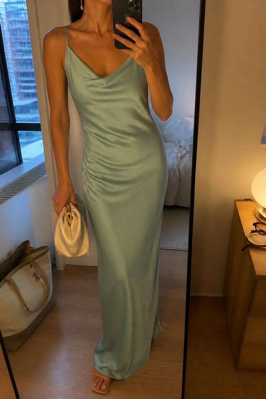 Image prompt
mirror selfie of a woman wearing a seafoam satin maxi dress with a soft drape, nude thin strap flat sandals, small gold drop earrings, and a small satin clutch, holding a phone in front of a mirror, cropped at NECK so face doesn't show, with a natural casual selfie pose, in a city apartment, warm moody lighting, including small lived-in details like a tote bag on the floor, sunglasses on a dresser, or soft bedding in the background, latest iPhone photo quality.
