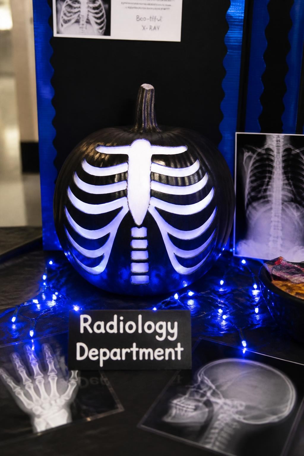 Image Prompt: A realistic DSLR photo of a black-painted pumpkin decorated with a glowing white skeleton ribcage design, soft blue LED lights around the base, mini x-ray film sheets leaning nearby, a small sign that says “Radiology Department,” clean dark display board, realistic hospital contest table setup, moody but family-friendly lighting.