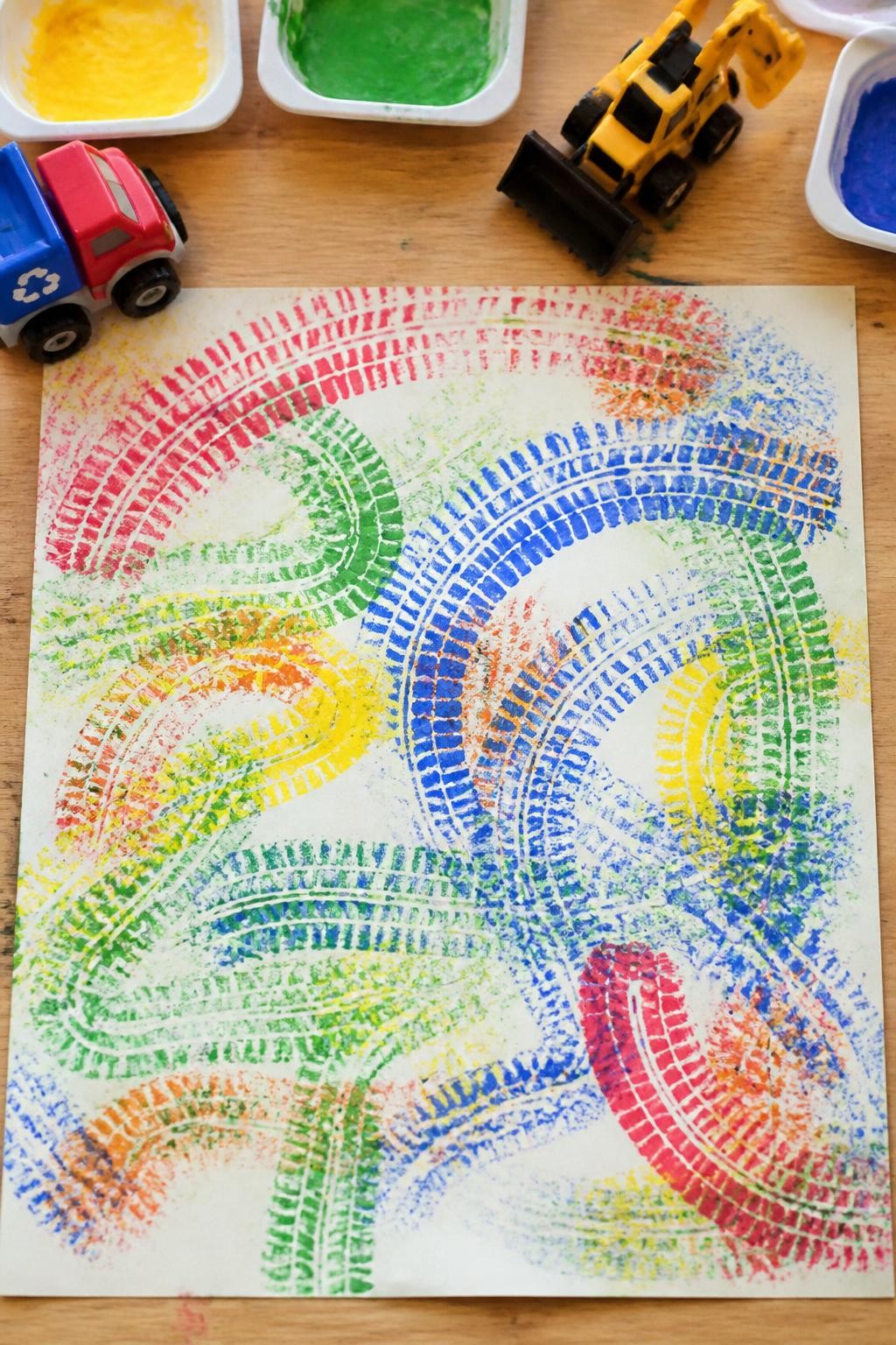 Image Prompt: Photo of finished work truck paint track art, overhead flat lay, placed on a wooden craft table with chunky toy trucks and shallow paint trays nearby, bright natural daylight, artwork filling most of the frame, bold tire marks and playful mixed colors clearly visible, realistic toddler craft photography, latest iPhone photo quality.