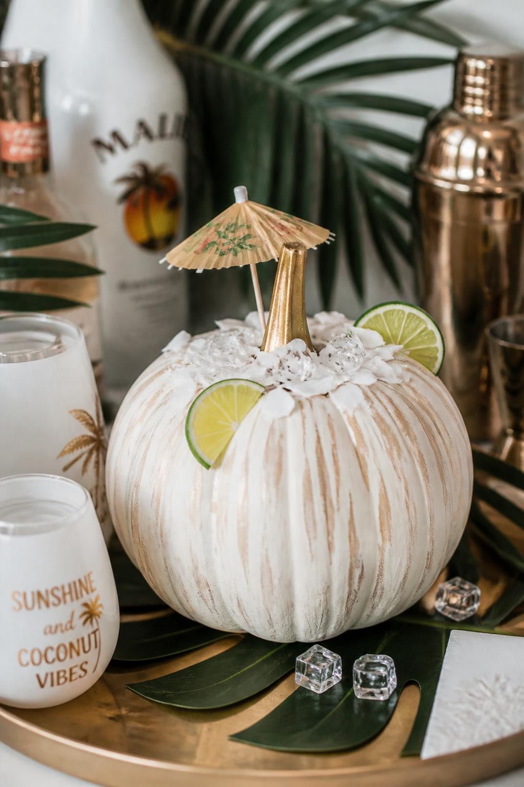 Image Prompt: A realistic DSLR photo of a creamy white pumpkin decorated with coconut-inspired texture, soft beige brush strokes, tiny lime wedge accents, faux coconut flakes around the stem, a small paper cocktail umbrella, and clear bead ice pieces, styled on a neutral bar tray with palm leaves and white glasses, elegant tropical fall decor.