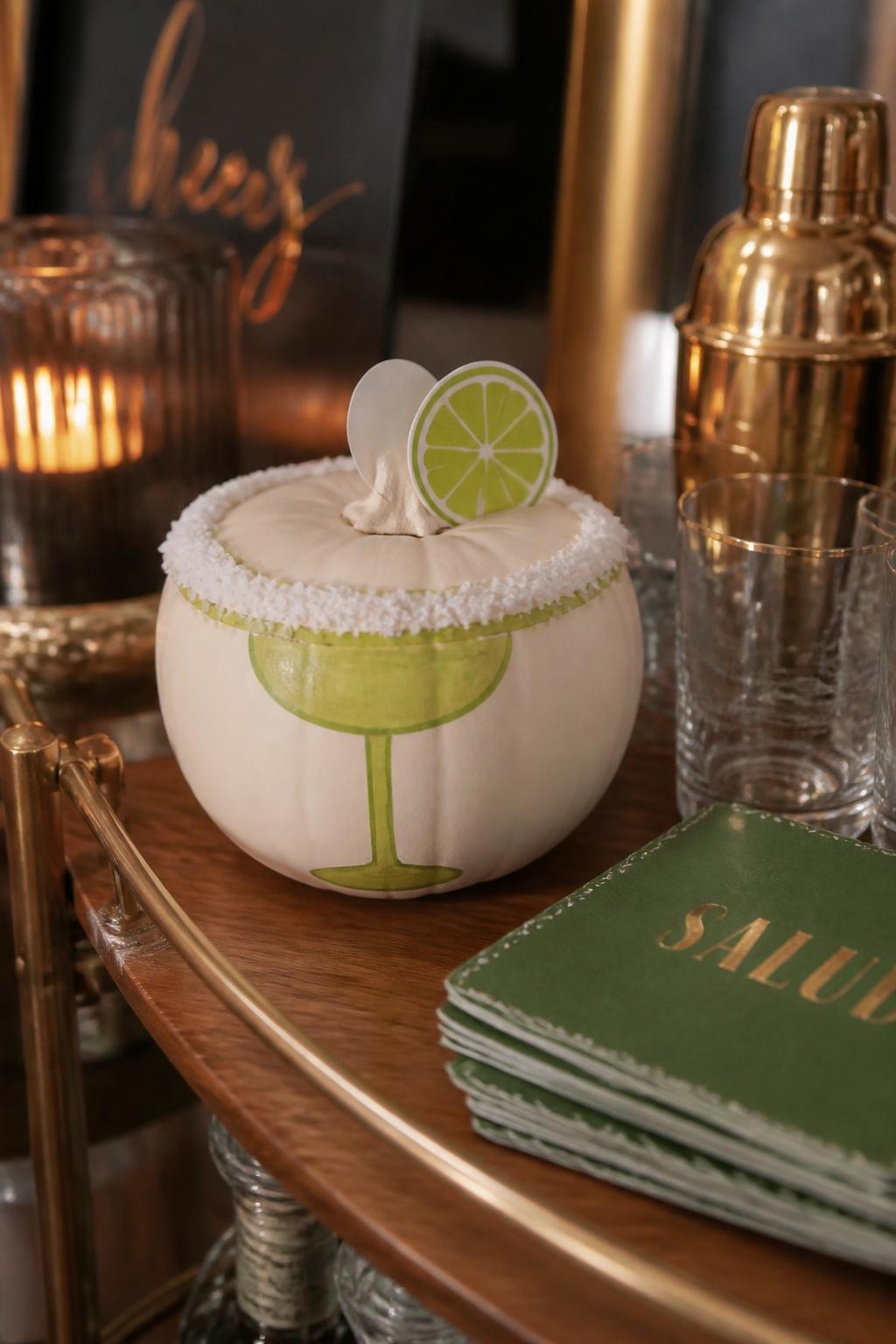 Image Prompt: A realistic DSLR photo of a small white pumpkin decorated like a margarita glass, with a thick textured faux salt rim around the top made from white craft crystals, lime green painted accents, a tiny paper lime wedge attached near the stem, placed on a wooden bar cart with cocktail napkins, clear glasses, and soft warm party lighting, stylish adult fall decor scene.