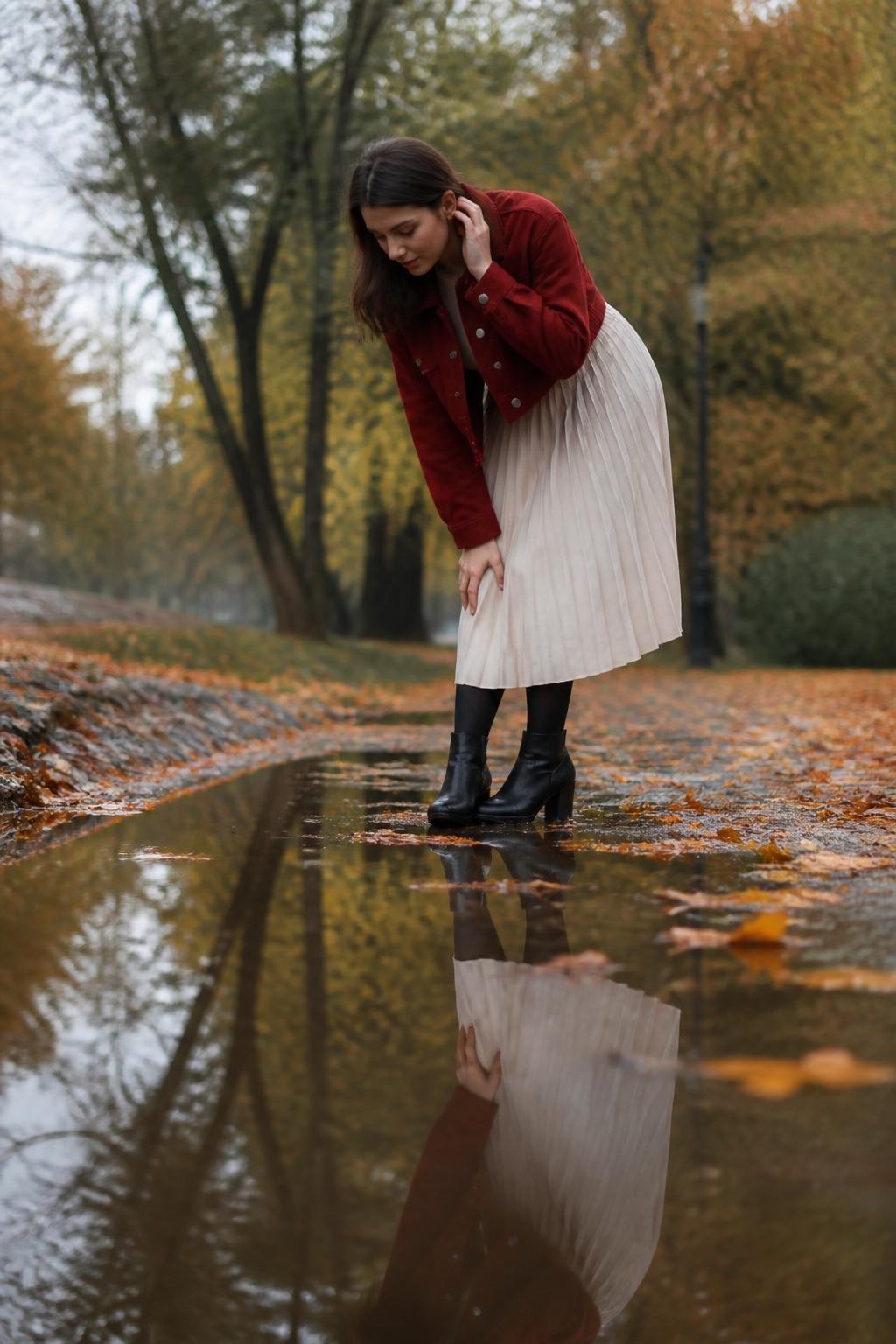 Image Prompt: A cinematic but realistic DSLR photograph taken after light fall rain, showing a young fair-skinned woman standing beside a large shallow puddle on a quiet paved path. She wears a cropped dark red jacket, a cream pleated midi skirt, black tights, and ankle boots. The camera is positioned low enough to capture her full reflection in the water. She bends slightly at the waist with one hand resting above her knee while the other tucks hair behind her ear. Her face turns toward the reflection rather than the lens. Wet orange leaves surround the puddle, blurred trees rise in the background, soft grey daylight, accurate water reflections, realistic skin and clothing texture, no artificial symmetry.