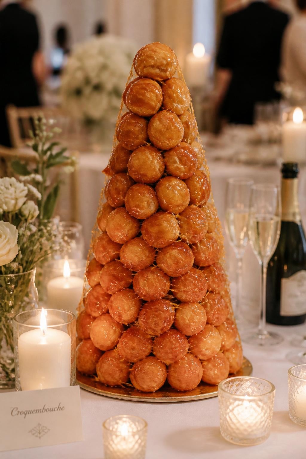 Image Prompt: Ultra-realistic wedding reception photo of a classic French croquembouche wedding cake made from stacked golden cream puffs with glossy caramel threads, arranged in a tall cone shape on an elegant dessert table. Place it in a real wedding party scene with candles, white flowers, fine linens, champagne glasses, French-inspired table styling, and a softly blurred reception backdrop. Keep the croquembouche as the clear main focus. No visible faces. If people appear, show only cropped hands, back views, side views, or neck-down figures. Realistic cream puff texture, caramel shine, warm romantic DSLR wedding photography. (no watermarks on images)