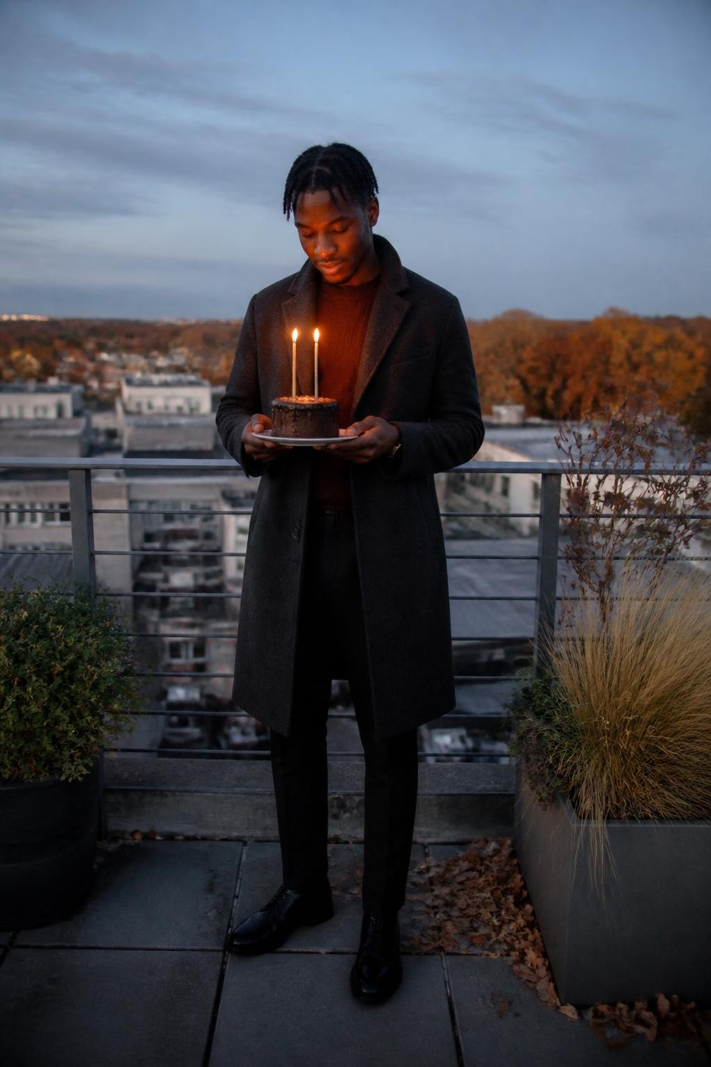 Image Prompt: A cinematic but realistic DSLR photograph of a young Black man standing on a secure rooftop terrace during late fall. He wears a long charcoal wool coat over a dark rust sweater, black tailored trousers, and polished boots. His hair is styled in neat short twists. He holds a small chocolate birthday cake on a sturdy ceramic plate, with two slim battery-operated candles creating a warm glow near his face. Low city buildings, rooftop plants, and distant amber trees sit beneath a pale blue evening sky. Natural dark brown skin tone, realistic low-light exposure, visible wool texture, secure railing, subtle wind movement, polished birthday fashion editorial.