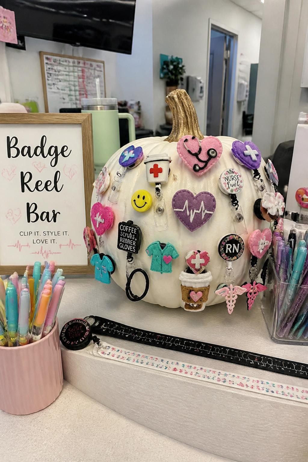 Image Prompt: A realistic DSLR photo of a white pumpkin covered with colorful handmade badge reels, mini felt hearts, tiny stethoscopes, smiley face stickers, coffee cup charms, and medical icons, displayed on a clean nurse station counter with pastel pens, lanyards, and a small sign reading “Badge Reel Bar,” trendy healthcare worker fall decor style.