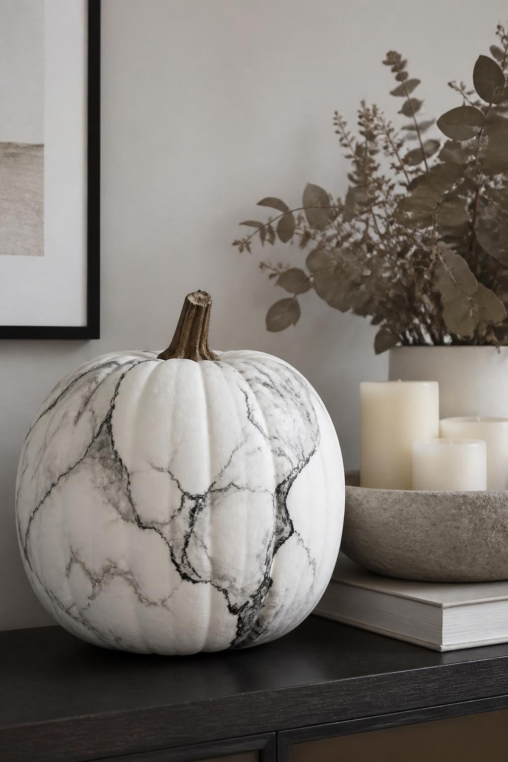 Horizontal 16:9 realistic iPhone photograph of a matte white real pumpkin painted with flowing black, charcoal, and pale-gray marble veins, irregular branching lines with selected edges gently blurred, natural brown stem, displayed on a modern console table beside a stone bowl, cream candles, neutral books, and dried eucalyptus, diffused daylight, realistic handmade marble effect, no artificial gloss, no text.