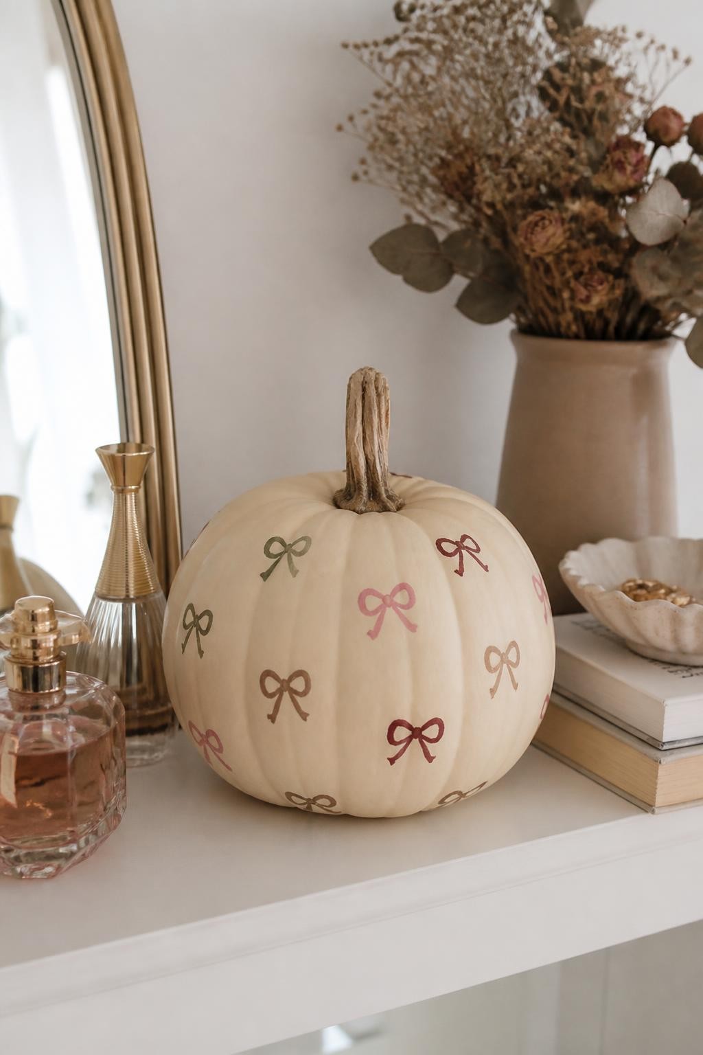 Horizontal 16:9 realistic iPhone photograph of a matte cream real pumpkin decorated with scattered tiny hand-painted bows in dusty rose, burgundy, muted sage, and soft brown, bows slightly varied in size and direction, natural twisted stem, displayed on a bedroom shelf beside perfume bottles, stacked books, dried flowers, and a ceramic dish, soft morning daylight, delicate handmade imperfections, cute polished fall decor, no text.