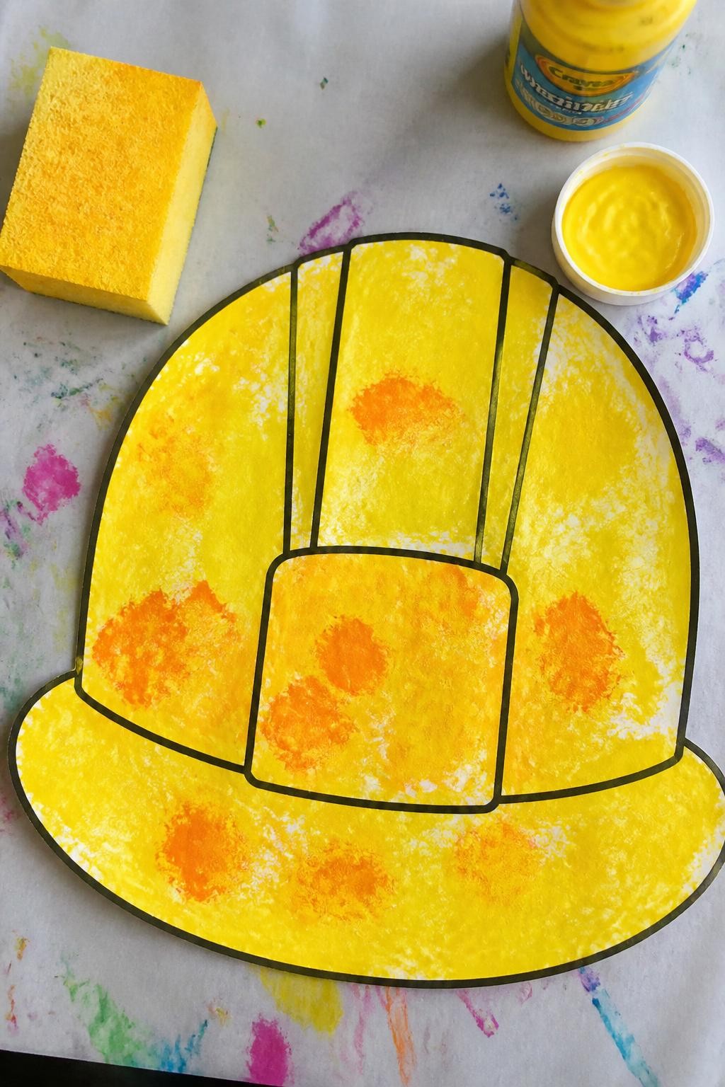 Image Prompt: Photo of a finished sponge-painted construction hard hat craft, overhead flat lay, placed on a colorful kids craft desk with a large sponge and washable yellow paint nearby, bright natural daylight, craft occupying most of the frame, chunky paint marks and simple toddler-made details clearly visible, realistic craft photography, latest iPhone photo quality.