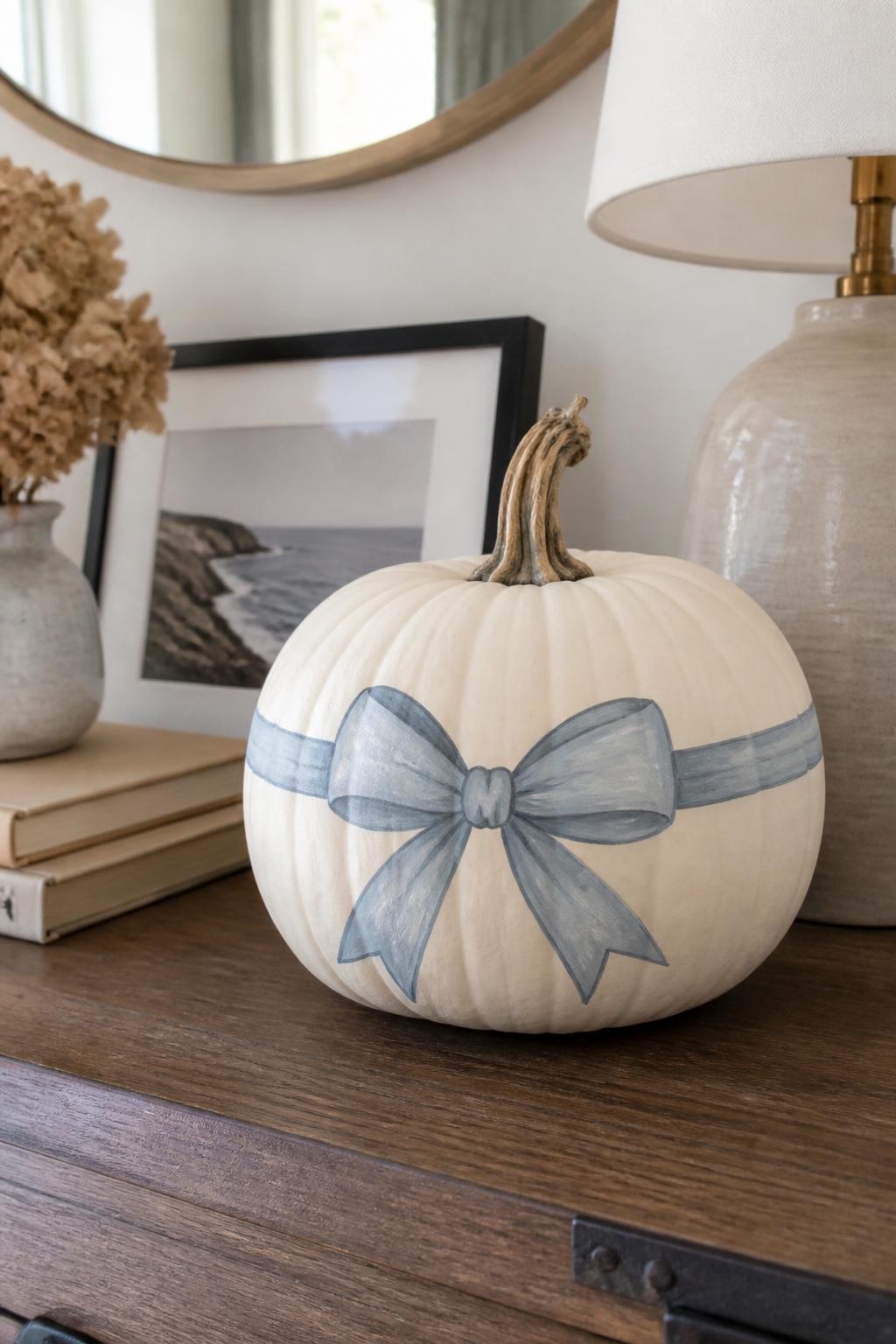 Horizontal 16:9 realistic iPhone photograph of a warm-white real pumpkin painted with a soft dusty-blue ribbon design wrapping around the widest section and forming a simple bow on the front, subtle taupe shading along the painted ribbon edges, natural twisted stem, displayed on an entryway console beside a ceramic lamp, framed photo, neutral books, and dried flowers, soft morning daylight, believable hand-painted fabric effect, no actual ribbon attached, no text.