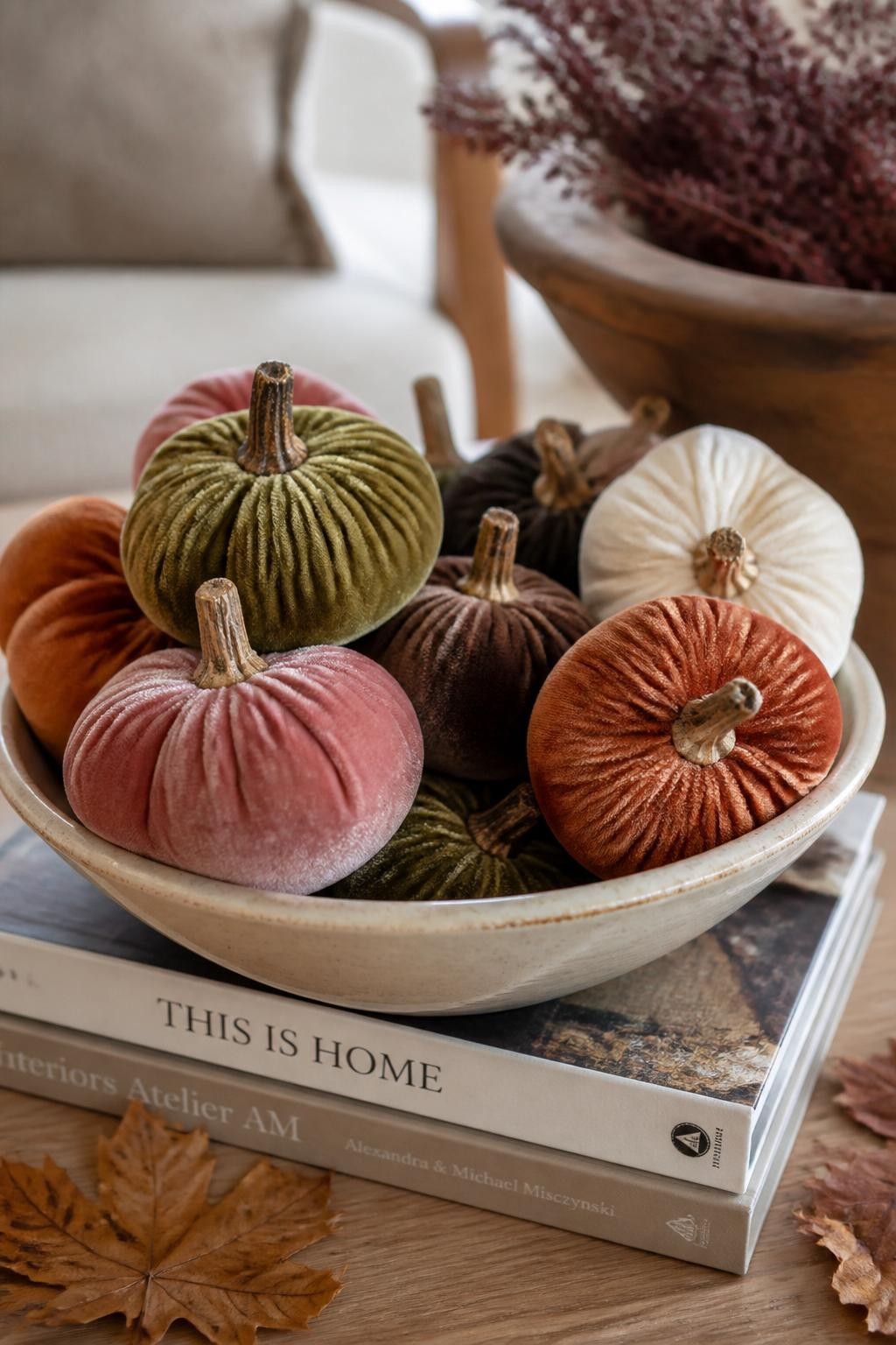 Image Prompt: A realistic DSLR close-up photo of small foam pumpkins wrapped in velvet fabric in olive green, dusty rose, rust, chocolate brown, and cream, with realistic brown stems, arranged in a shallow ceramic bowl on a coffee table with books, dried leaves, and warm autumn daylight, cozy elegant handmade fall decor style.