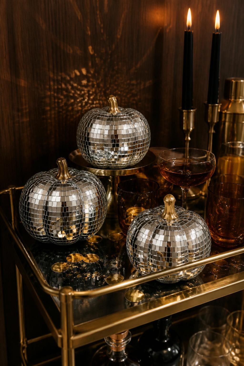 Image Prompt: A realistic close-up DSLR photo of small pumpkins covered in tiny mirrored disco tiles, sitting on a modern fall bar cart with amber glasses, black taper candles, metallic confetti, and a dark wood backdrop, warm evening light, slight sparkle reflection on the wall, trendy party decor style.