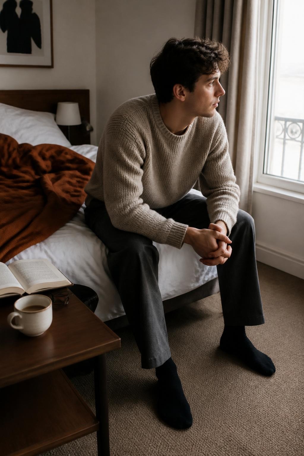 Image Prompt: A realistic DSLR lifestyle image of a young fair-skinned man sitting on the edge of an unmade bed in a softly lit hotel-style bedroom. He wears a textured oatmeal sweater, loose charcoal trousers, and dark socks. A rust-colored duvet is folded unevenly across white bedding, while an open paperback, reading glasses, and a half-finished cup of coffee rest on the bedside table. He leans forward with his elbows on his knees, looking toward the window. Pale morning light, subtle shadow under the bed, realistic fabric creases, natural posture, restrained autumn palette, intimate editorial atmosphere.