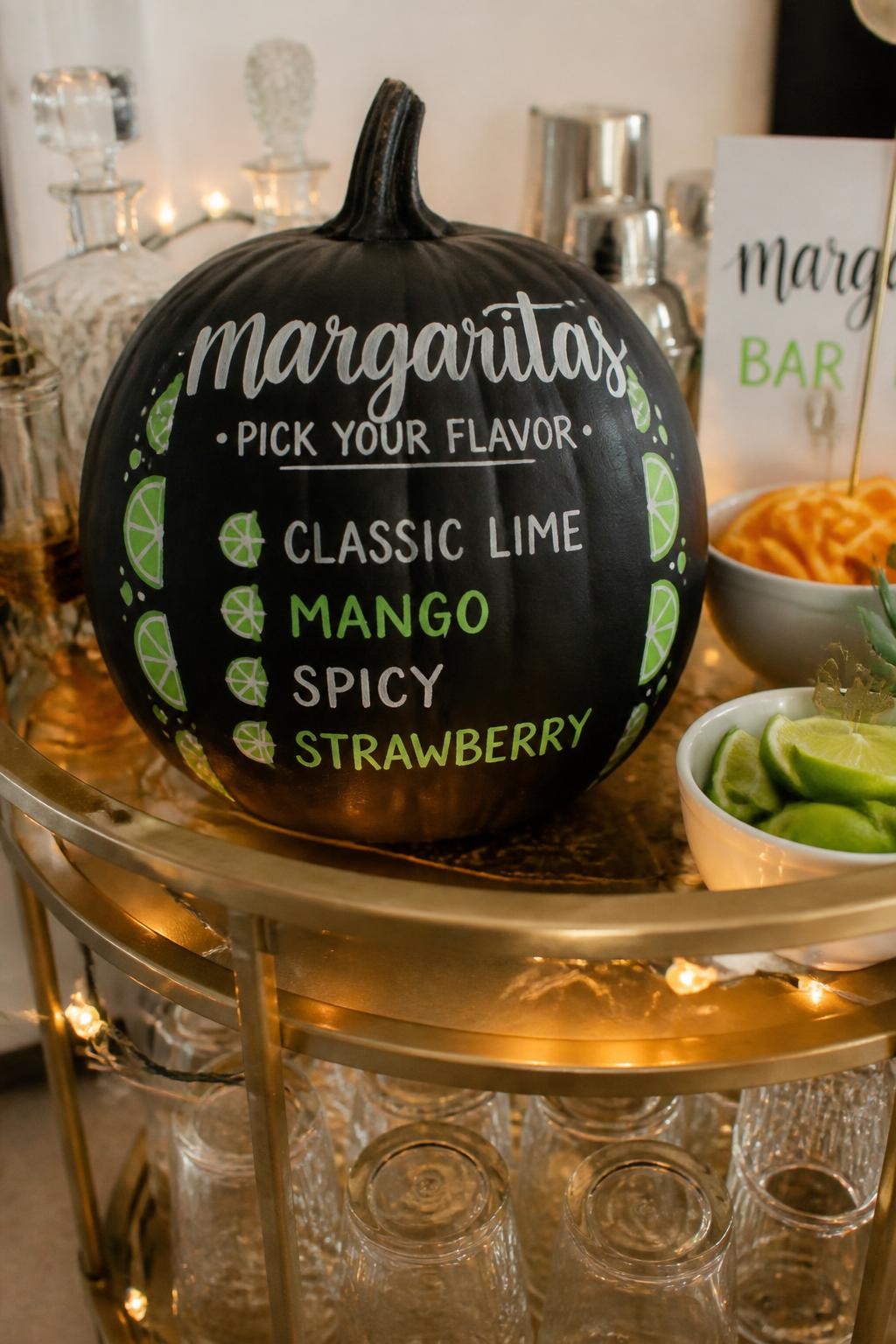 Image Prompt: A realistic DSLR photo of a medium matte black pumpkin used as a mini margarita bar menu, with hand-lettered white and lime green text listing flavors like classic, mango, spicy, and strawberry, small painted lime border, styled on a bar cart with glasses, citrus bowls, and warm string lights, modern party signage photography.