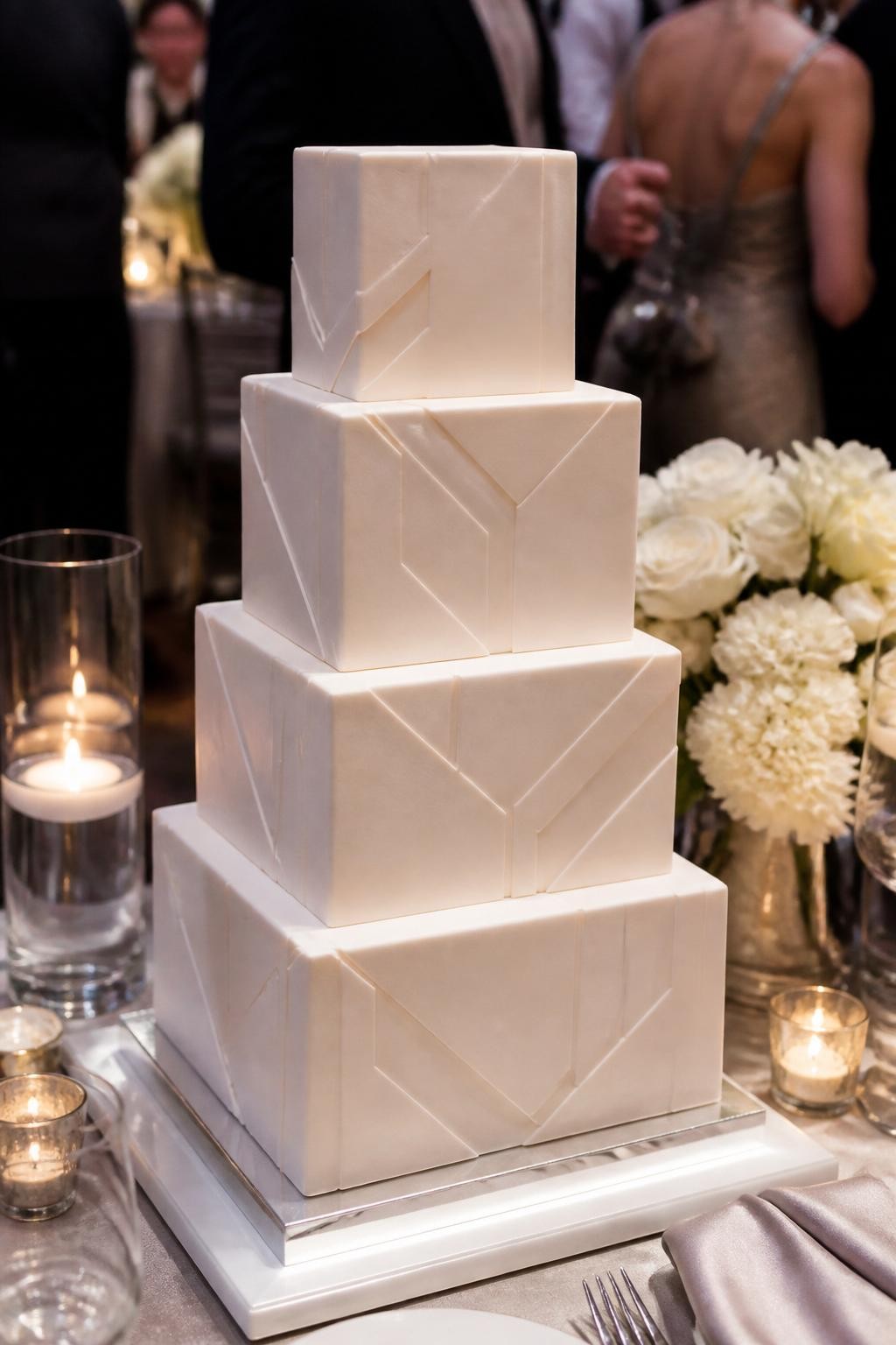 Image Prompt: Ultra-realistic wedding party image featuring a modern geometric square wedding cake with sharp tiers, raised geometric panels, clean lines, and subtle tone-on-tone details in white, ivory, or soft beige. Place the cake on a sleek reception cake table with candles, modern florals, glassware, neutral linens, and a softly blurred venue backdrop. Keep the cake as the hero of the photo. No visible faces. If guests appear, show only cropped torsos, hands, back views, or side views in blur. Realistic fondant texture, crisp geometric detail, soft DSLR event lighting. (no watermarks on images)