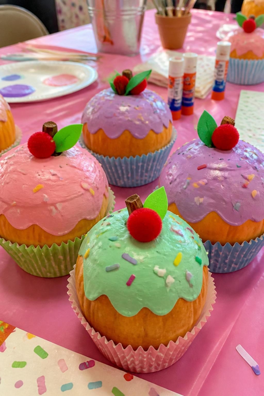 Image Prompt: A realistic DSLR photo of small pumpkins painted like cupcakes with pastel tops, paper baking cup bases, colorful sprinkle stickers, tiny pom-pom cherries, and craft foam frosting shapes, arranged on a bright kids party table with paint trays, glue sticks, and napkins, cheerful no-carve pumpkin decorating scene, realistic handmade details.