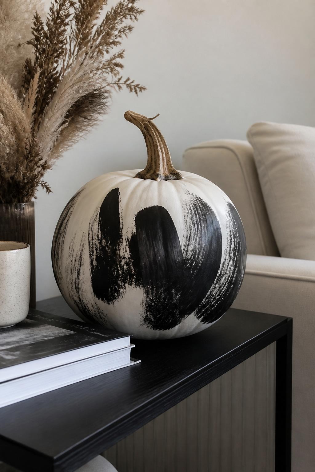 Horizontal 16:9 natural iPhone photo of a matte white real pumpkin decorated with broad expressive black brushstrokes across one side, visible bristle marks, faded edges, small white spaces between strokes, natural twisted stem, displayed on a modern side table beside art books, a ceramic cup, and dried grasses, soft diffused daylight, believable handmade artwork, no perfect digital shapes, no text.