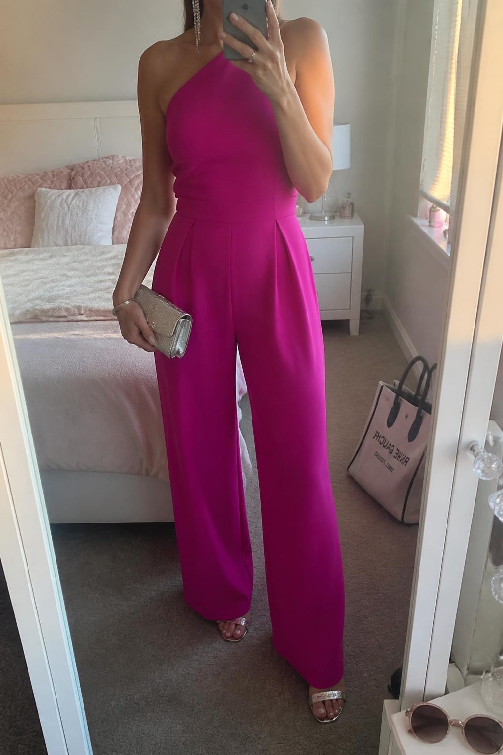 Image prompt
mirror selfie of a woman wearing a fuchsia wide-leg jumpsuit with silver heeled sandals, a metallic silver clutch, and crystal drop earrings, holding a phone in front of a mirror, cropped at NECK so face doesn't show, with a natural casual selfie pose, in a feminine-themed bedroom, evening golden hour, including small lived-in details like sunglasses on a dresser and a tote bag on the floor, latest iPhone photo quality.