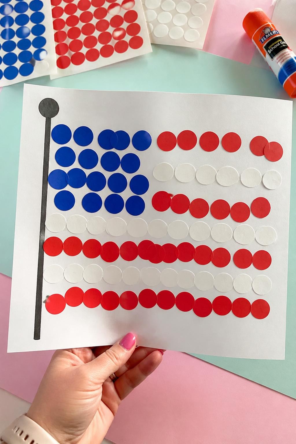 Image Prompt: Photo of a finished dot sticker Labor Day flag craft, held in hand POV, above a pastel craft mat workspace with large sticker sheets and a glue stick nearby, bright natural lighting, flag filling most of the frame, oversized dots and uneven toddler placement clearly visible, realistic craft photography, latest iPhone photo quality.