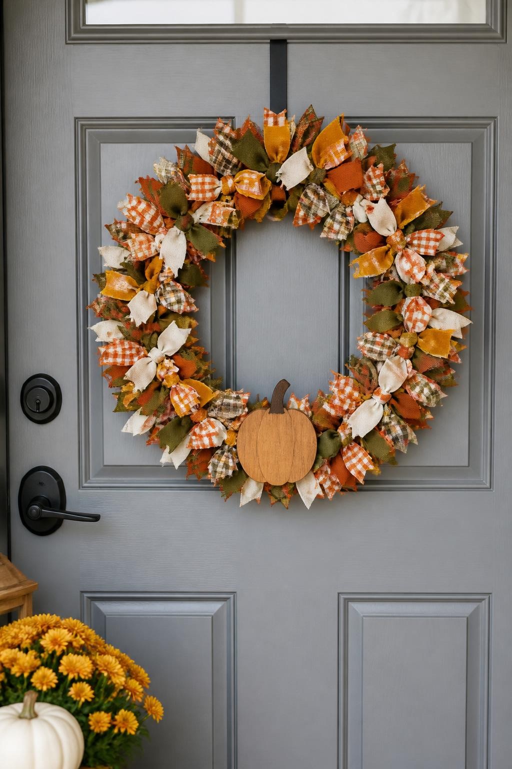 Image prompt: A cheerful handmade fall wreath hanging on a warm gray front door, created from hundreds of short ribbon loops tied closely around a wire frame, using rust gingham, mustard velvet, cream linen, olive cotton, brown plaid, and muted orange grosgrain ribbon, arranged in balanced sections rather than random clutter, finished with a small wooden pumpkin at the bottom, realistic fabric textures and neatly trimmed ends, bright but soft daylight, family-friendly porch, DSLR photography, no people, vertical format.