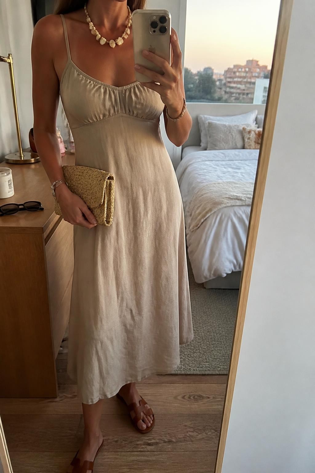 mirror selfie of a woman wearing a soft neutral midi dress, beachy statement necklace, tan flat leather sandals, and a small straw clutch, holding a phone in front of a mirror, cropped at NECK so face doesn't show, with a natural casual selfie pose, in a city apartment, evening golden hour, including small lived-in details like sunglasses on a dresser, or soft bedding in the background, latest iPhone photo quality.