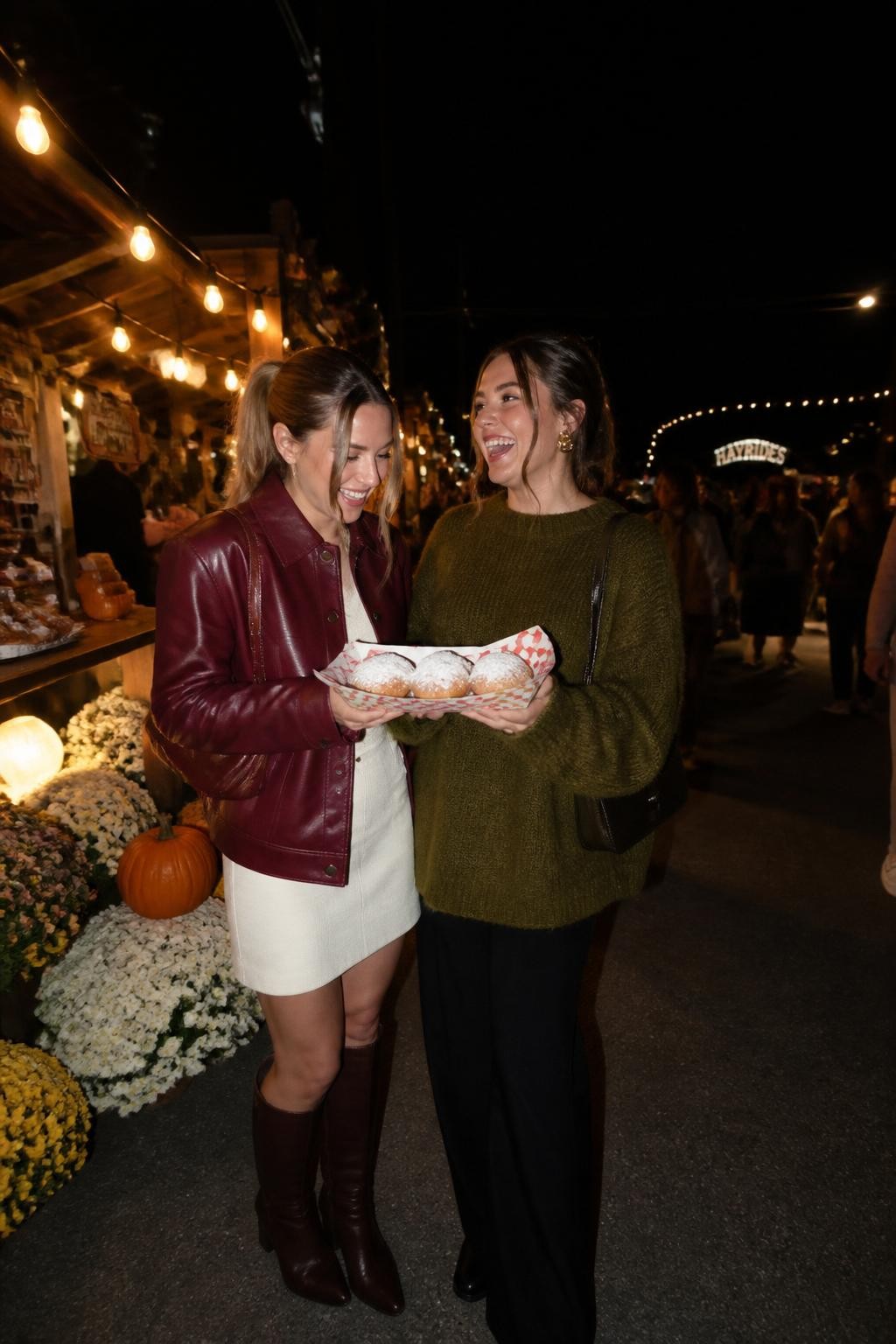 Image Prompt: A realistic nighttime flash photograph of two female friends at an outdoor fall market with string lights, wooden stalls, chrysanthemums, baked goods, and people moving in the background. One woman wears a dark cherry leather jacket with a cream skirt and boots, while the other wears a moss green sweater with relaxed black trousers. They stand close together holding a paper tray of pastries, caught laughing rather than posing directly. Strong direct-camera flash creates sharp foreground detail while the background remains darker with glowing lights and slight motion blur. Authentic nightlife photography, realistic expressions, crisp fabric texture, casual 2026 Instagram style.