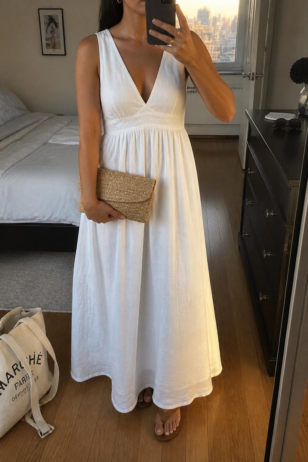 Image prompt
mirror selfie of a woman wearing a white linen blend maxi dress with a flowy skirt, tan flat leather sandals, small gold hoop earrings, and a woven straw clutch with a clean shape, holding a phone in front of a mirror, cropped at NECK so face doesn't show, with a natural casual selfie pose, in a city apartment, evening golden hour, including small lived-in details like a tote bag on the floor, sunglasses on a dresser, or soft bedding in the background, latest iPhone photo quality.