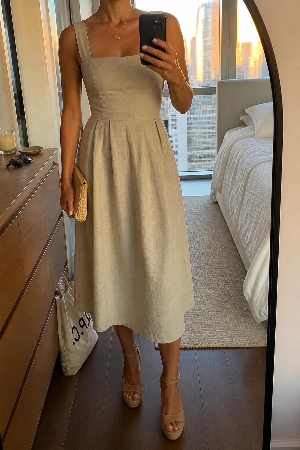 Image prompt
mirror selfie of a woman wearing a sand linen blend midi dress with a tailored waist, nude wedge espadrilles, a delicate gold pendant necklace, and a small straw clutch, holding a phone in front of a mirror, cropped at NECK so face doesn't show, with a natural casual selfie pose, in a city apartment, evening golden hour, including small lived-in details like a tote bag on the floor, sunglasses on a dresser, or soft bedding in the background, latest iPhone photo quality.