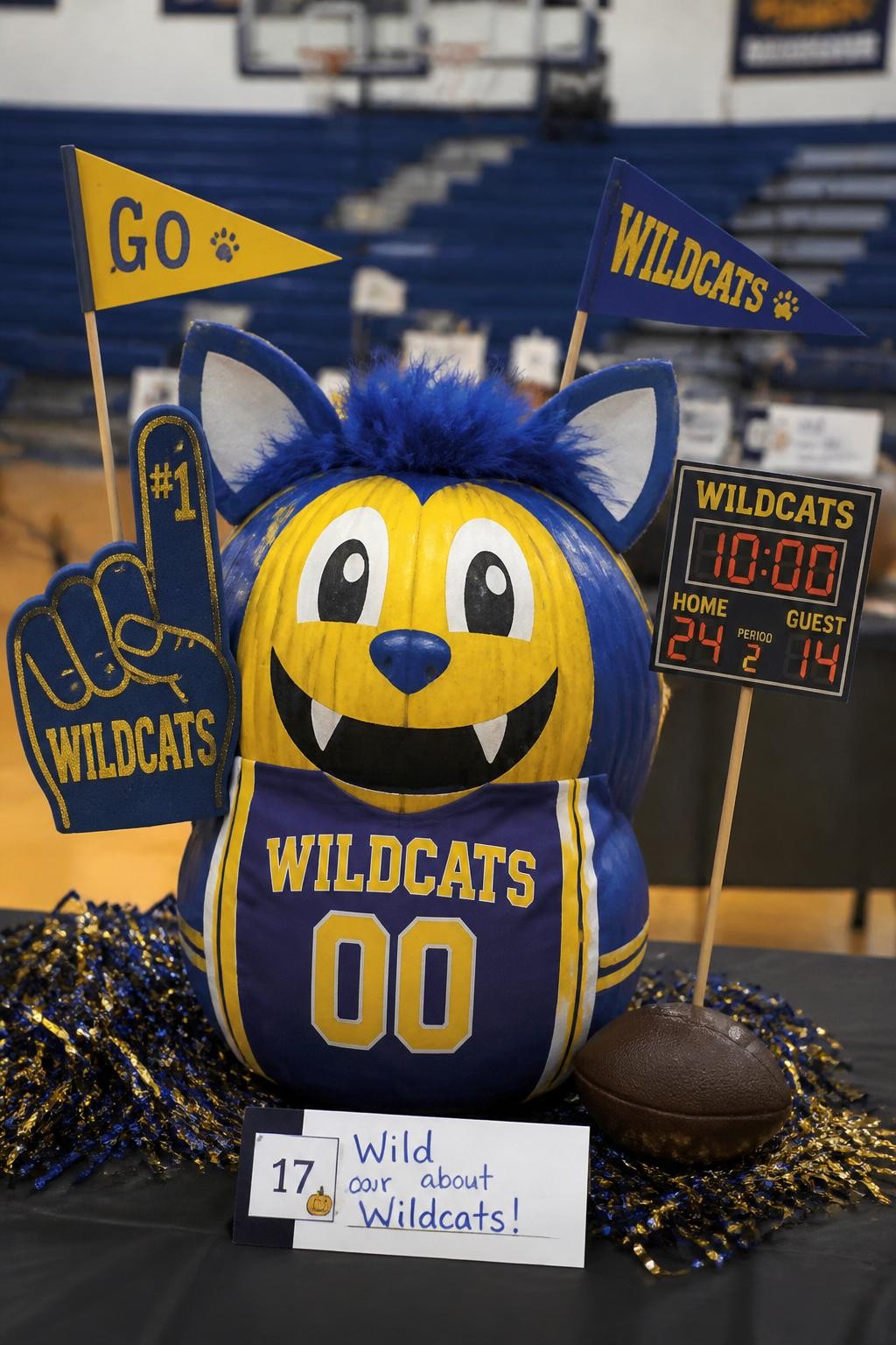 Image Prompt: A realistic DSLR photo of a pumpkin decorated as a school mascot, with school-color paint, paper jersey, felt ears or mascot features, foam finger prop, mini pennant flags, and a small scoreboard sign, displayed on a gym table with blurred bleachers in the background, bright indoor school spirit lighting, festive contest display.