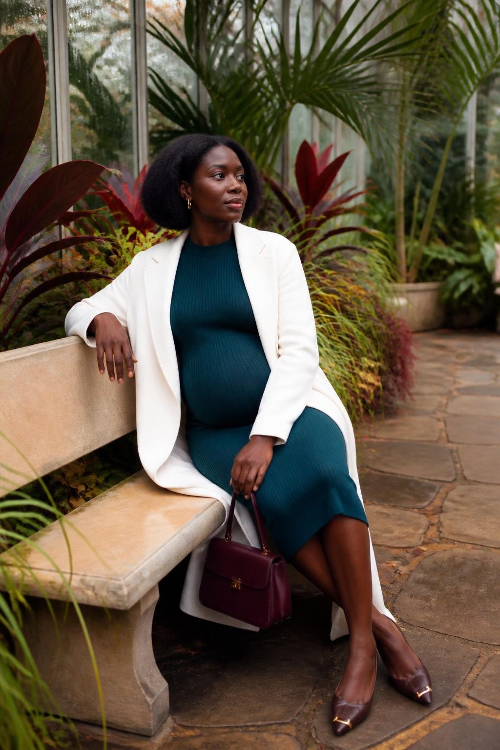 Image Prompt: A realistic DSLR maternity-free solo portrait of a young Black woman seated on a pale stone bench inside a botanical conservatory during fall. She has deep brown skin and wears a fitted dark teal midi dress beneath a long cream coat, pointed brown flats, and slim gold earrings. Her natural hair is styled in a polished rounded bob. One arm rests along the bench back while the other hand holds a small structured burgundy bag near her knee. Large burgundy leaves, deep green palms, warm stone paths, and condensation-covered glass surround her. She sits with both knees angled to one side and her shoulders turned gently toward the light. Accurate deep skin tone, realistic plant and fabric textures, diffused daylight, graceful body proportions, sophisticated botanical editorial.