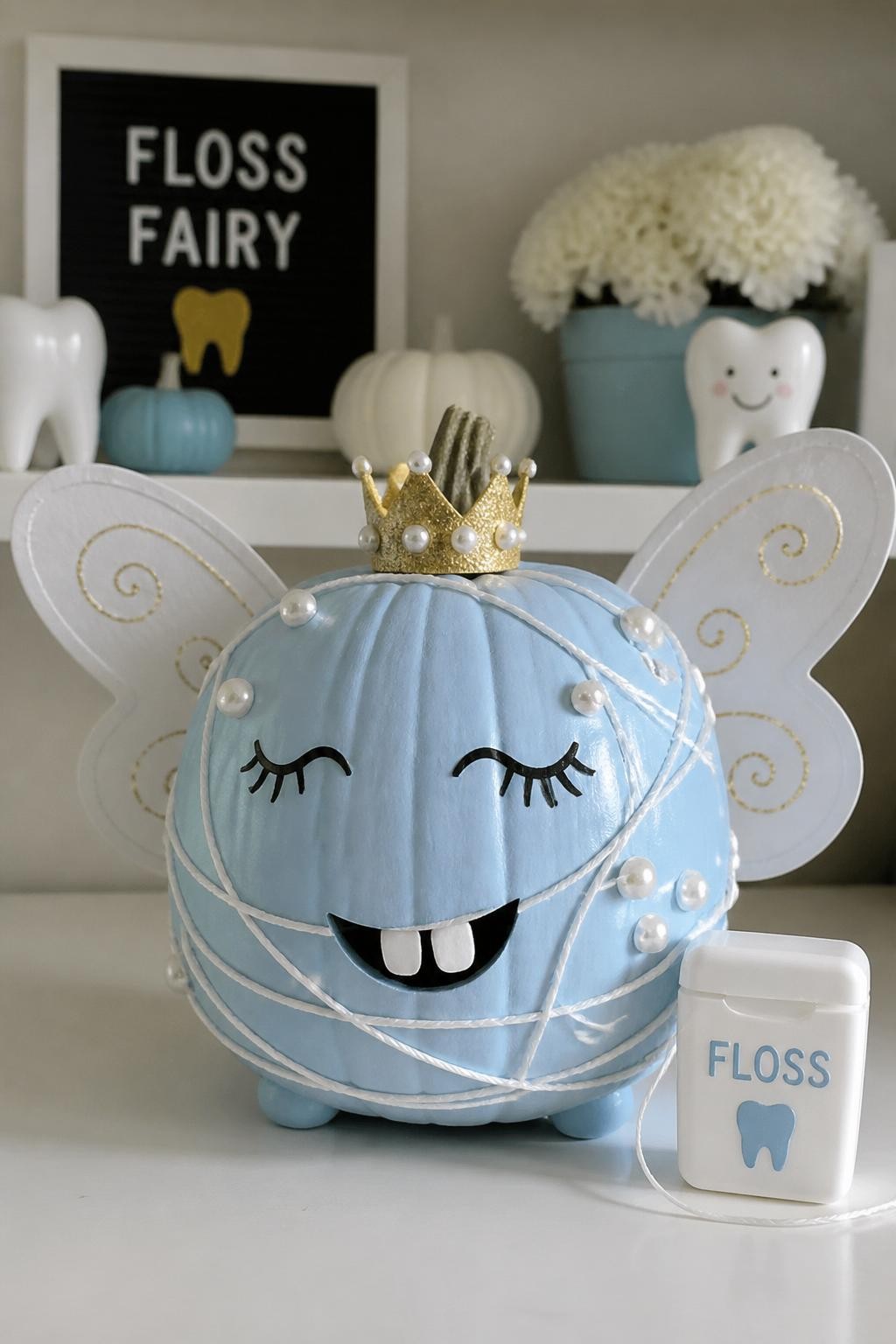 Image Prompt: A realistic DSLR photo of a pale blue pumpkin decorated as a floss fairy, with white string swirls wrapped gently around the pumpkin, paper wings, a tiny crown, painted lashes, small tooth cutouts, pearl stickers, and a floss container prop beside it, displayed on a clean white craft table with soft daylight, cute dental-themed fall decor.