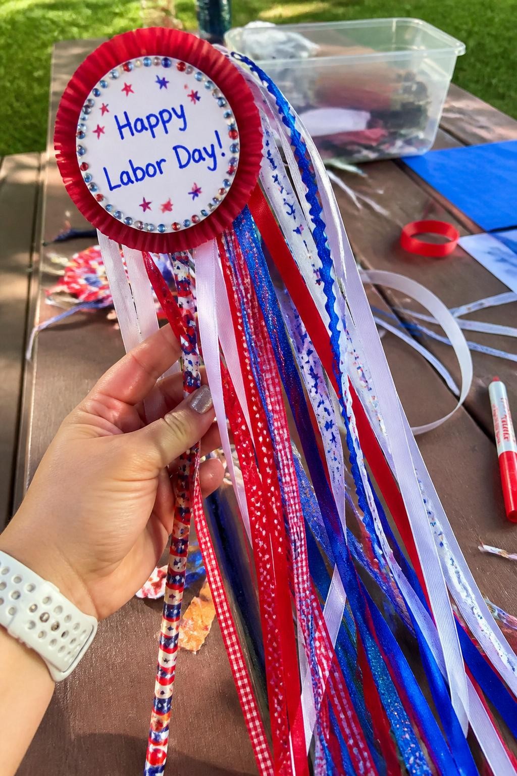 Image Prompt: Photo of a finished Labor Day ribbon parade wand, held in hand POV, above an outdoor picnic table craft activity with ribbon scraps and a washable marker nearby, bright natural daylight, wand and flowing ribbons occupying most of the frame, colorful handmade details clearly visible, realistic craft photography, latest iPhone photo quality.