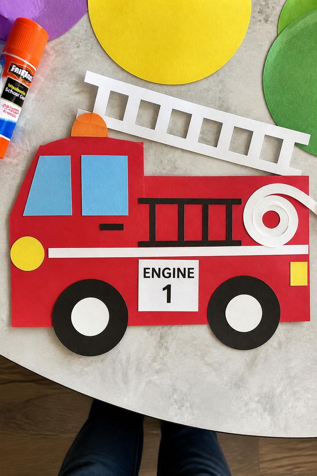 Image Prompt: Photo of a finished shape-built fire truck collage, overhead flat lay, arranged on a colorful kids craft desk with large paper circles and a glue stick nearby, bright natural daylight, fire truck occupying most of the frame, bold shapes and uneven toddler placement clearly visible, realistic craft photography, latest iPhone photo quality.