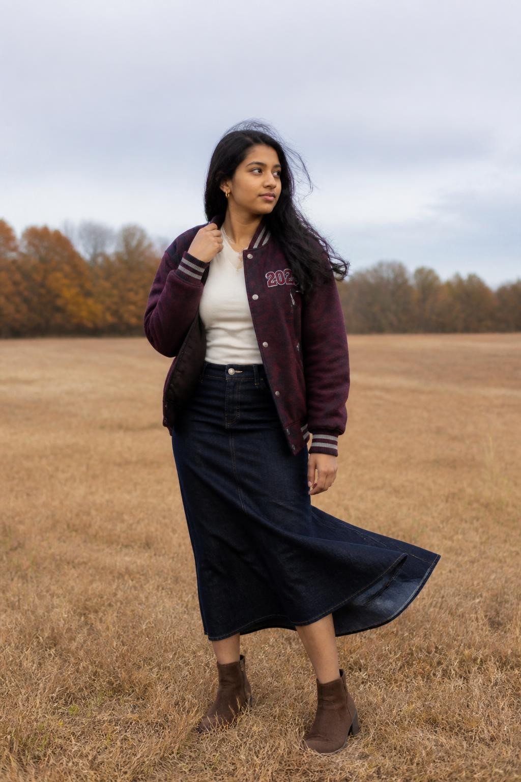 Image Prompt: A realistic full-body DSLR photograph of a South Asian teenage girl standing in an open field of faded golden grass on a windy fall afternoon. She wears a deep burgundy varsity jacket customized with a small graduation-year patch, a fitted cream top, a dark denim midi skirt, and brown ankle boots. She holds the jacket collar lightly with one hand while the other hangs naturally at her side. Her long dark hair and skirt move gently in the wind as she looks across the field. Low amber trees and a pale grey sky create a minimal background. Natural brown skin texture, realistic jacket stitching, soft directional light, subtle movement, contemporary senior fashion portrait.