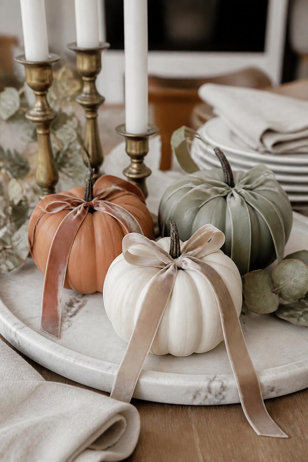 Image Prompt: A realistic DSLR photo of small matte pumpkins in cream, terracotta, and sage green with long velvet ribbons tied around their stems, placed on a marble tray with brass candle holders, dried eucalyptus, and folded linen napkins, soft neutral fall decor, elegant home styling, natural daylight.