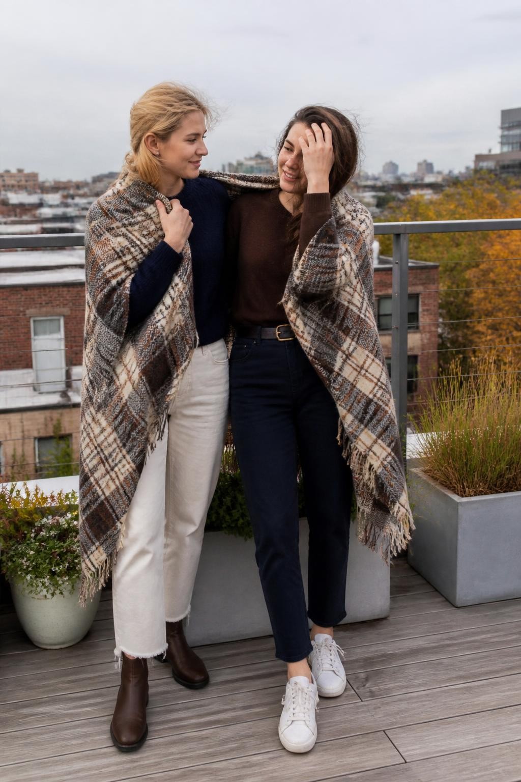 Image Prompt: A realistic full-body DSLR photograph of two young women on a safe low-rise rooftop terrace during a breezy fall afternoon. They stand side by side wrapped loosely in one oversized checked wool blanket, wearing simple outfits in navy, chocolate, and cream underneath. One friend holds the blanket closed near her shoulder while the other reaches up to keep her hair from blowing across her face. Low city buildings, rooftop plants, and amber trees appear in the background. Cool natural light, realistic wind movement, detailed wool texture, secure visible terrace railing, candid expressions, contemporary city lifestyle photography.